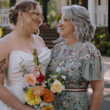 Load image into Gallery viewer, Two women standing together outdoors, holding flowers, with a building in the background.